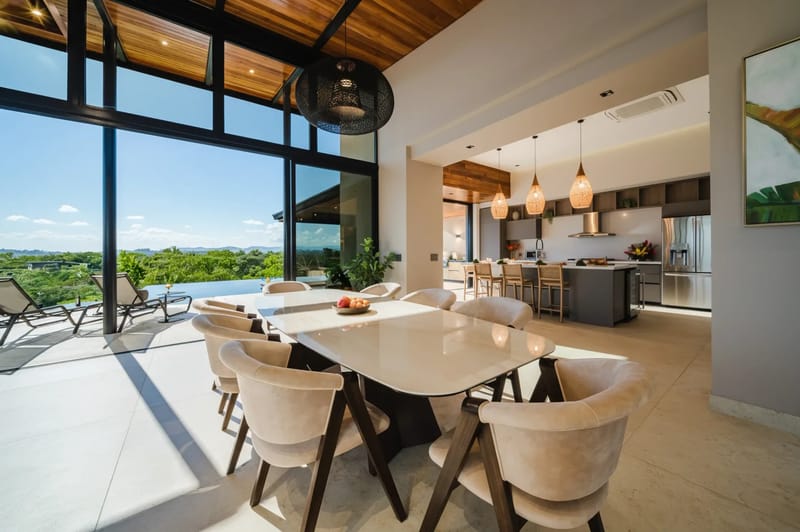 Casa Ama · 7. The bright dining area, featuring a large table and seating for eight under a wood-paneled ceiling, opens through expansive glass walls to a tropical landscape view and an outdoor pool, seamlessly connecting to a modern kitchen with island seating.