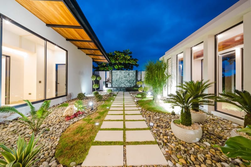 Casa Ama · 51. A modern villa entrance features an illuminated pathway through tropical gardens, flanked by glass walls revealing warm interiors under a twilight sky.