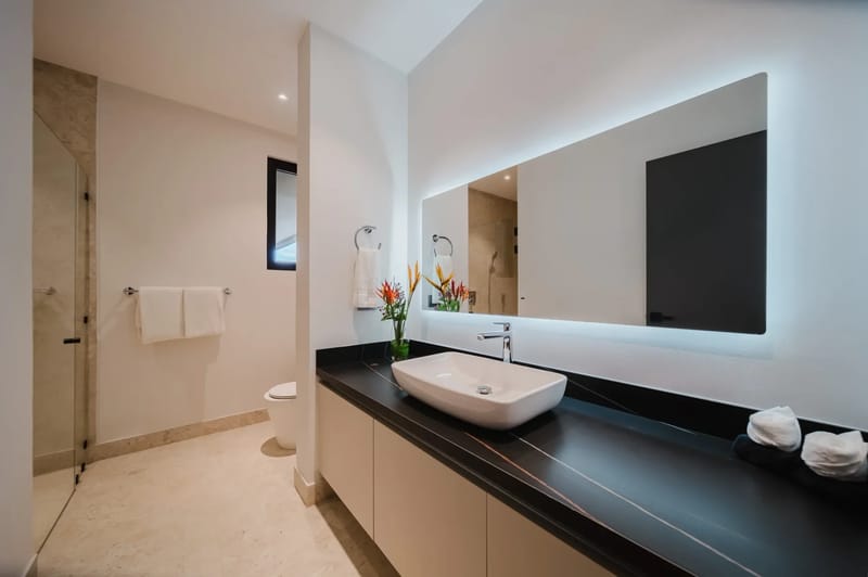 Casa Ama · 46. A bright, modern bathroom features a sleek vanity with a vessel sink, a large backlit mirror, and a walk-in shower with a glass enclosure.