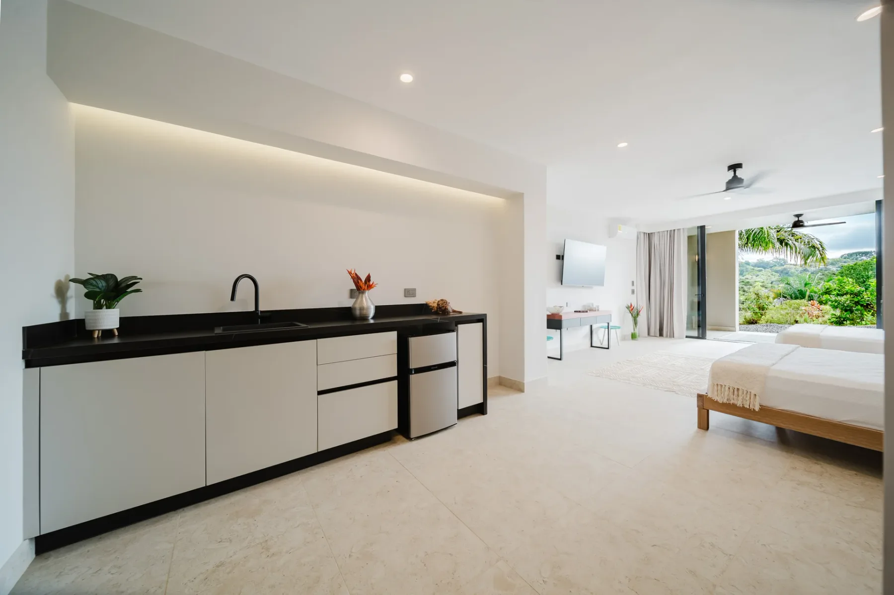Casa Ama · 14. This bright bedroom suite includes two beds and a small kitchenette, with wide glass doors opening to a lush tropical garden and ample natural light.