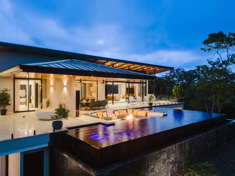 Casa Ama · 30. This modern villa at dusk features illuminated indoor living spaces opening onto a multi-level infinity pool and terrace, set against lush tropical vegetation.