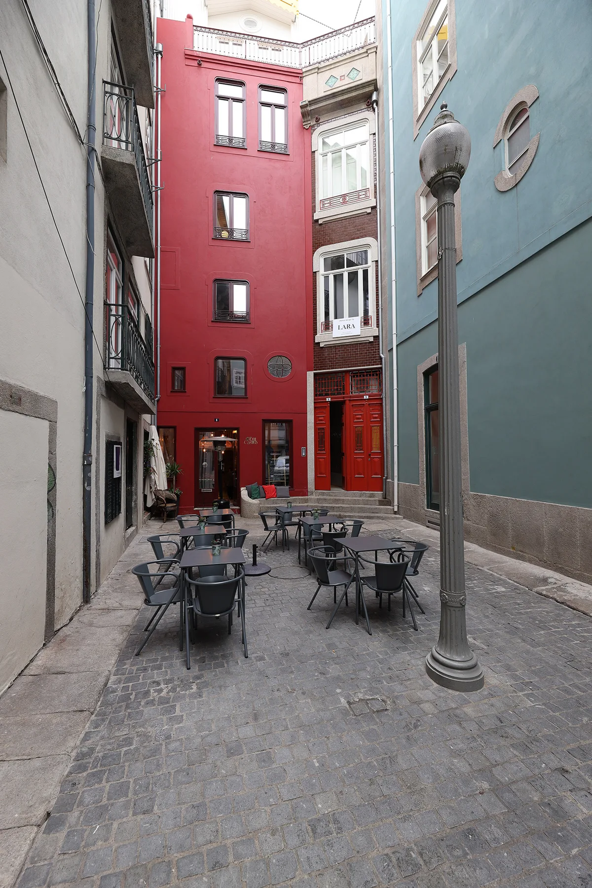 Un patio estrecho y pavimentado entre edificios coloridos tiene mesas y sillas al aire libre.
