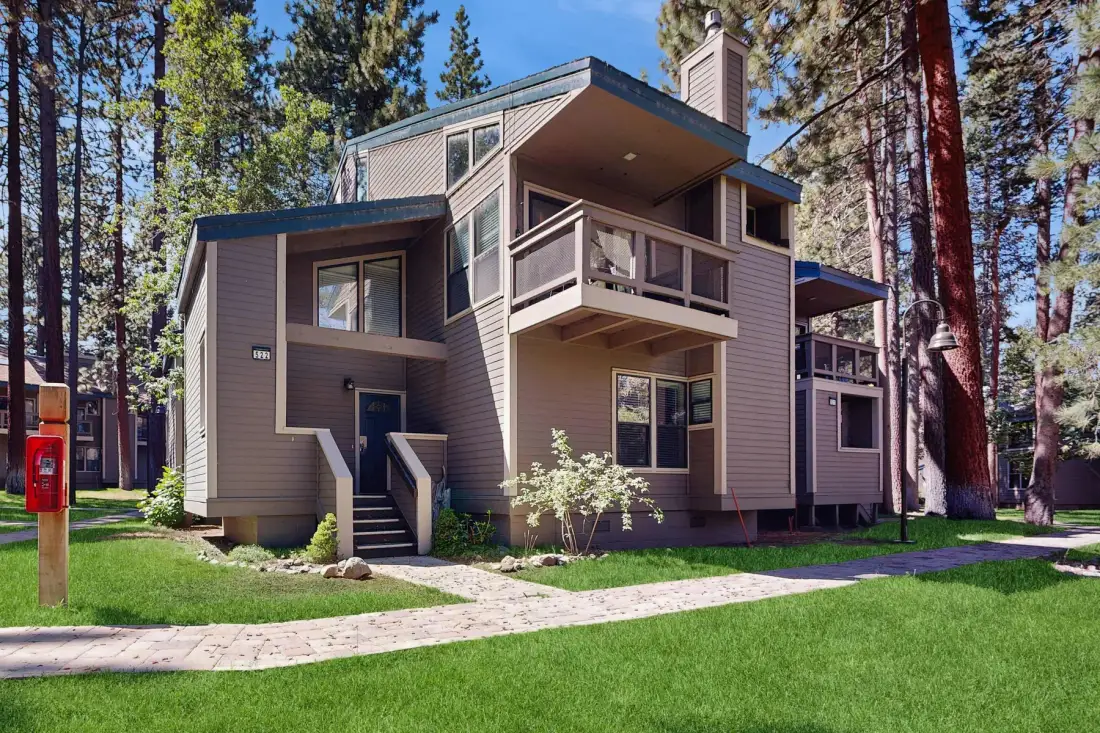 A modern multi-level house stands among tall trees beside a green lawn.