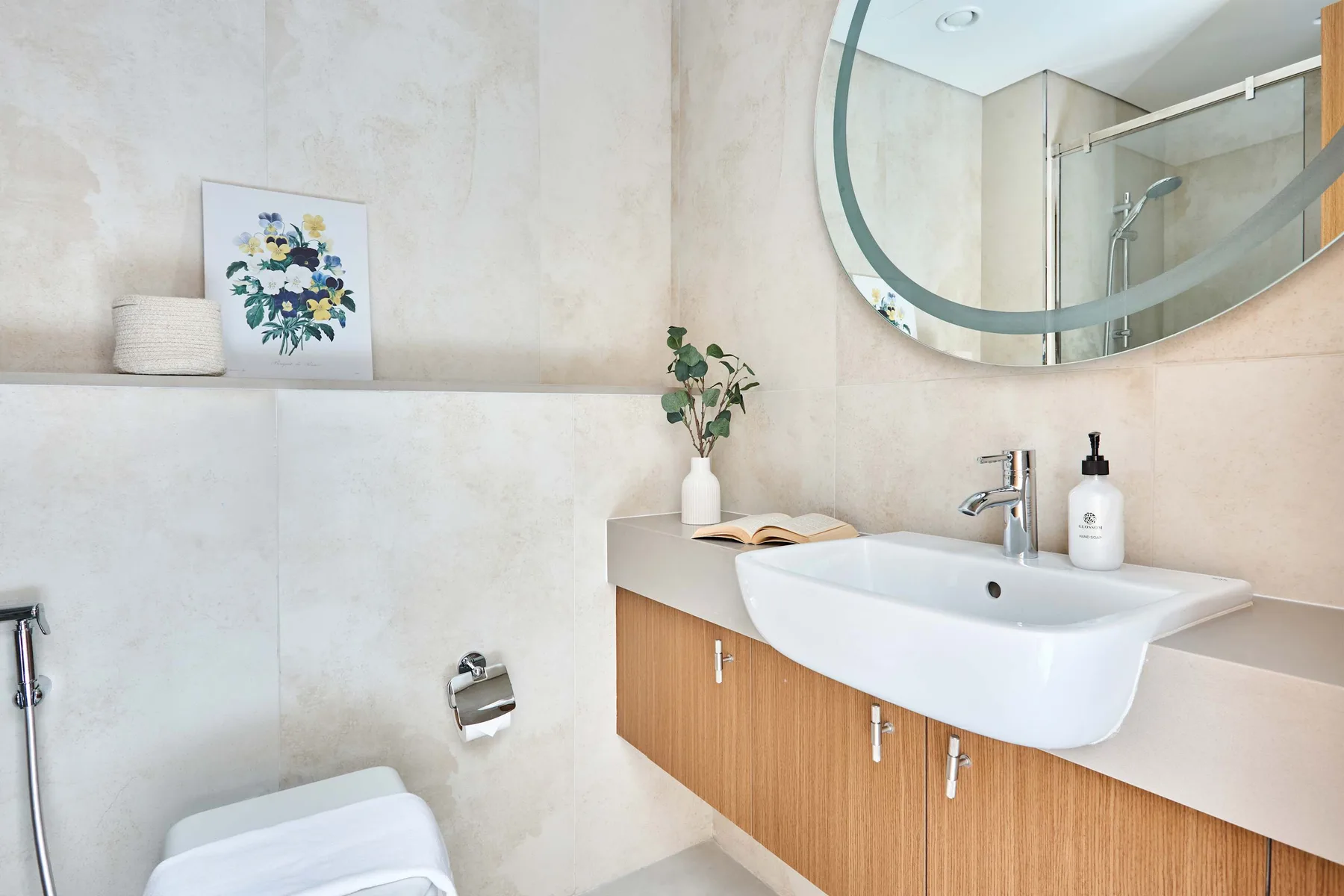 Un baño luminoso con lavabo blanco, muebles de madera y un gran espejo redondo.