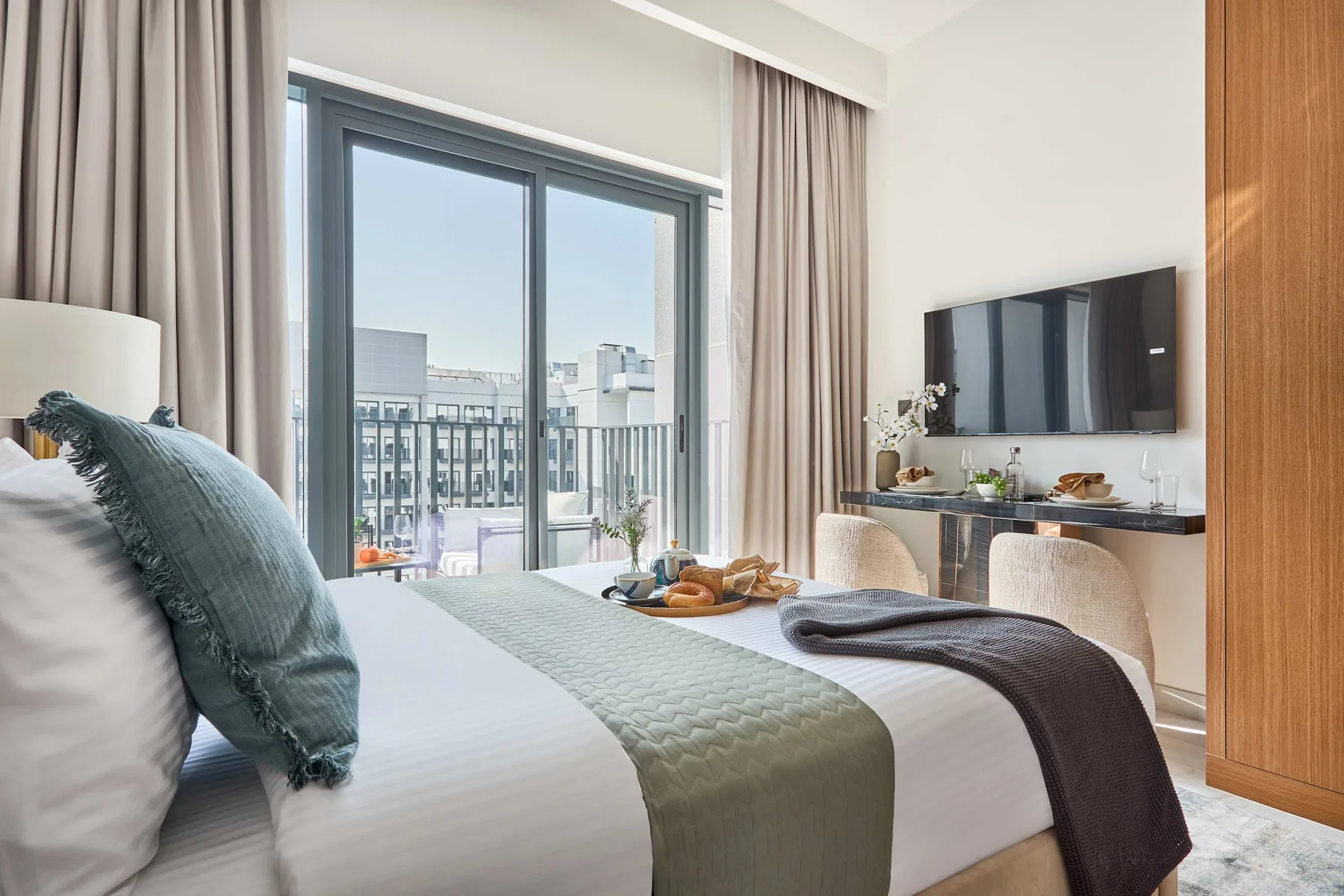 Un dormitorio luminoso con una gran ventana, una cama tendida y un televisor montado en la pared.