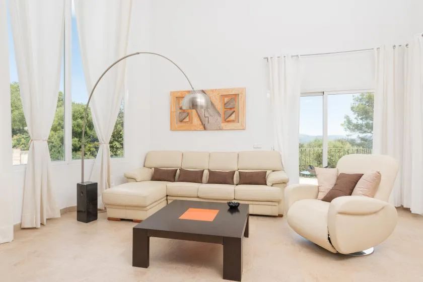 Una sala luminosa con asientos beige, una mesa de centro oscura y ventanas altas con cortinas.