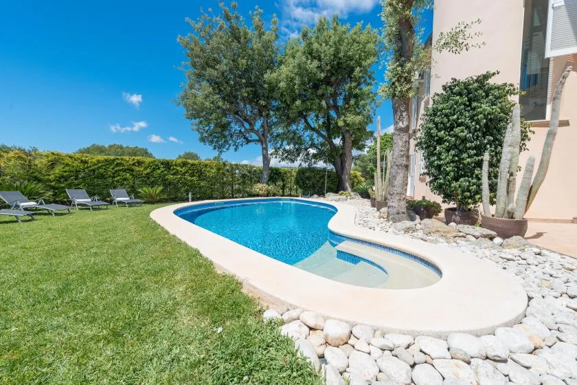 Una piscina al aire libre está junto a un césped, árboles y un edificio de color claro.