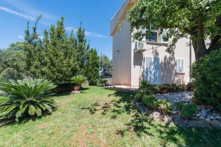 Un jardín junto a una casa clara, con árboles, arbustos y un arriate bordeado de piedras.