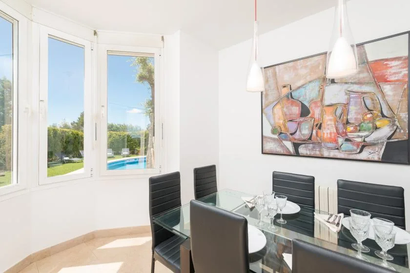 Un comedor luminoso tiene una mesa de vidrio junto a grandes ventanas con vistas al jardín.