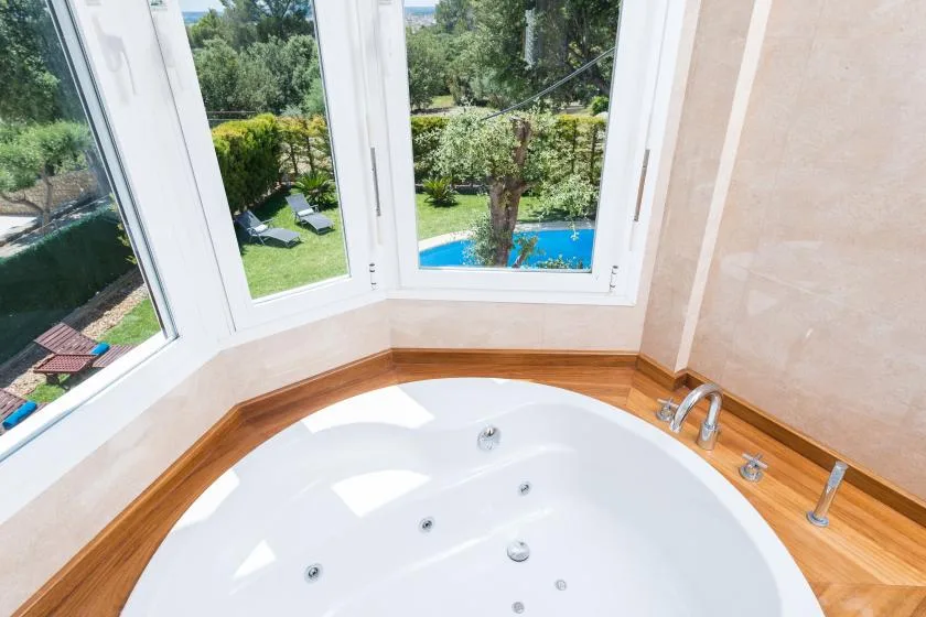 Una bañera está junto a grandes ventanas con vistas a un jardín y una piscina.