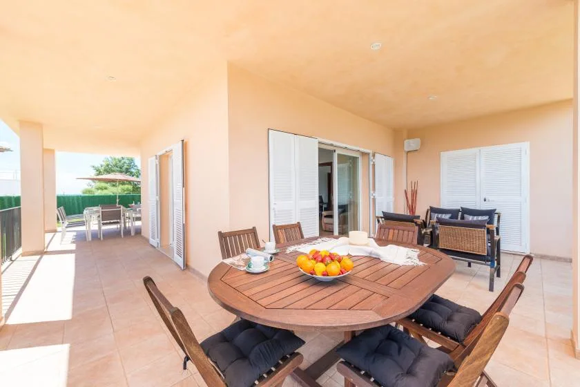 Un patio cubierto tiene una mesa de comedor de madera y asientos junto a puertas corredizas abiertas.