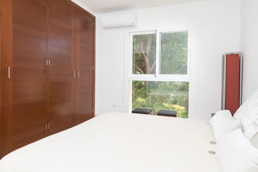 Un dormitorio luminoso con una cama, armarios de madera y una ventana con vistas a la vegetación.