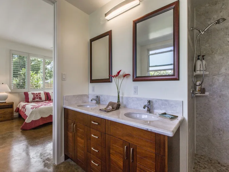 Un tocador de baño con dos lavabos está junto a una ducha de vidrio y una puerta abierta al dormitorio.