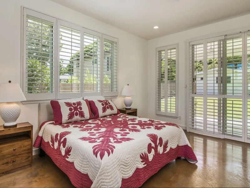 Un dormitorio con una cama cubierta por una colcha estampada roja y blanca junto a ventanas con persianas.