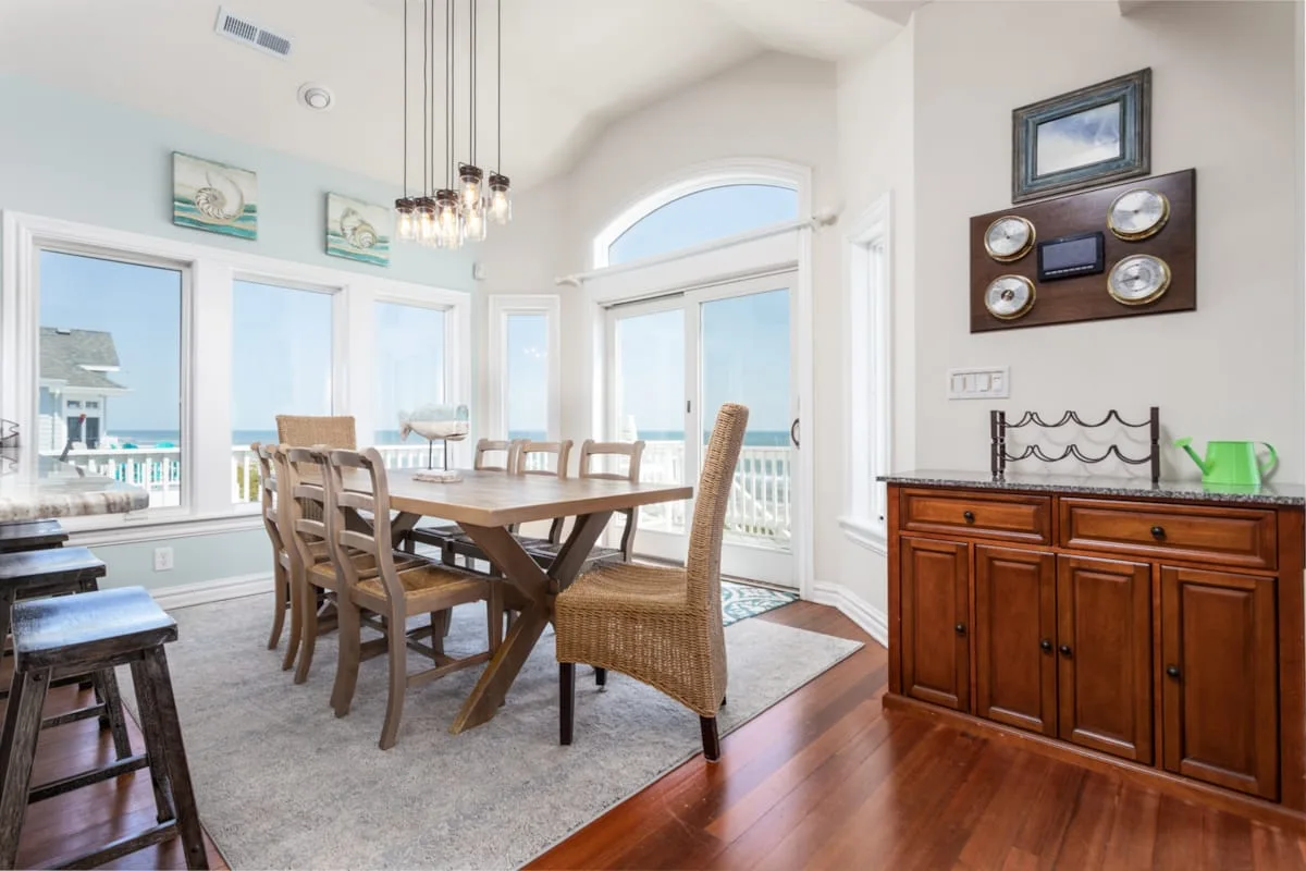 A bright dining room with a long wooden table and large windows overlooking the water.