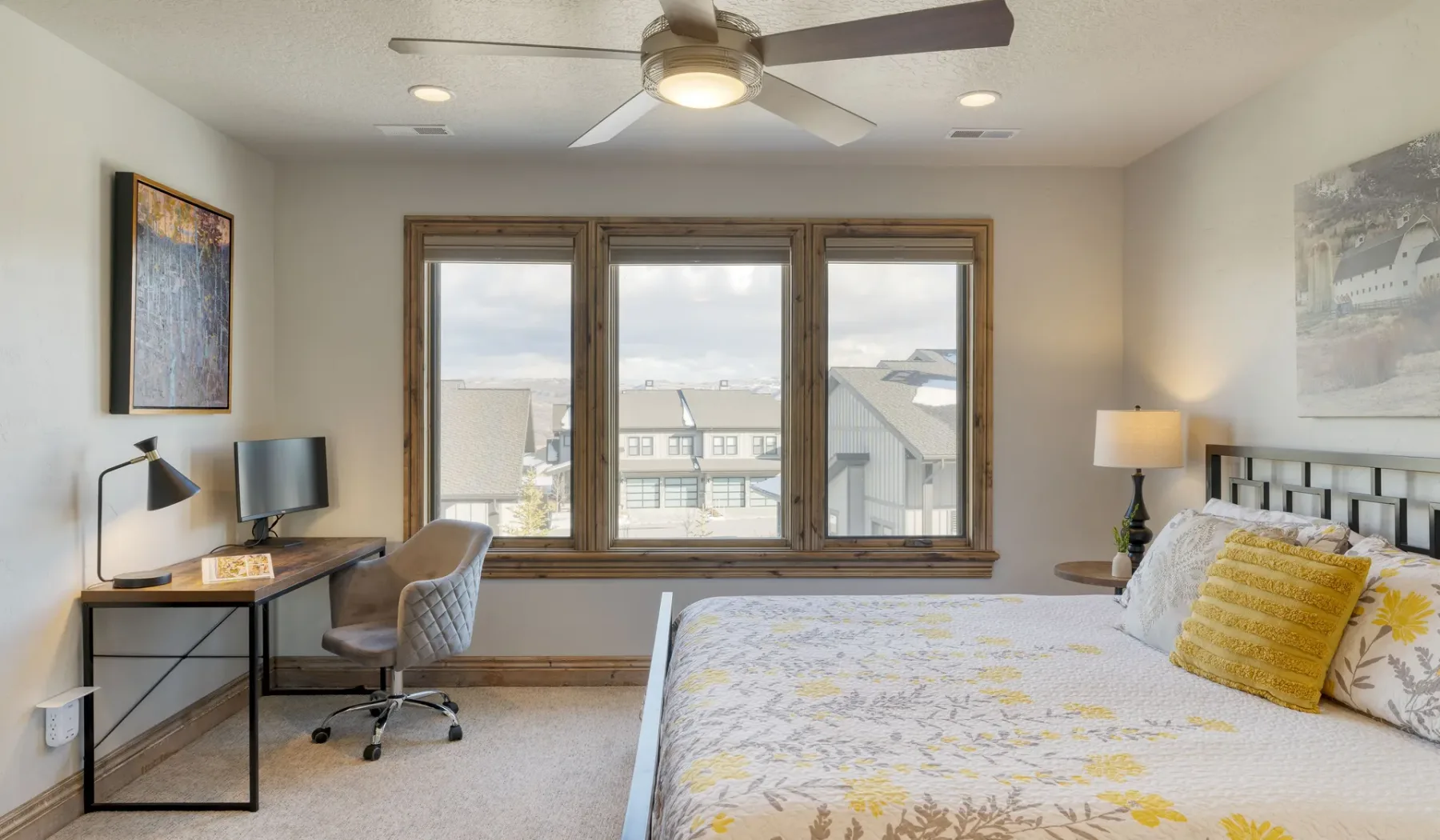 A bedroom with a bed, desk, and windows overlooking nearby buildings.
