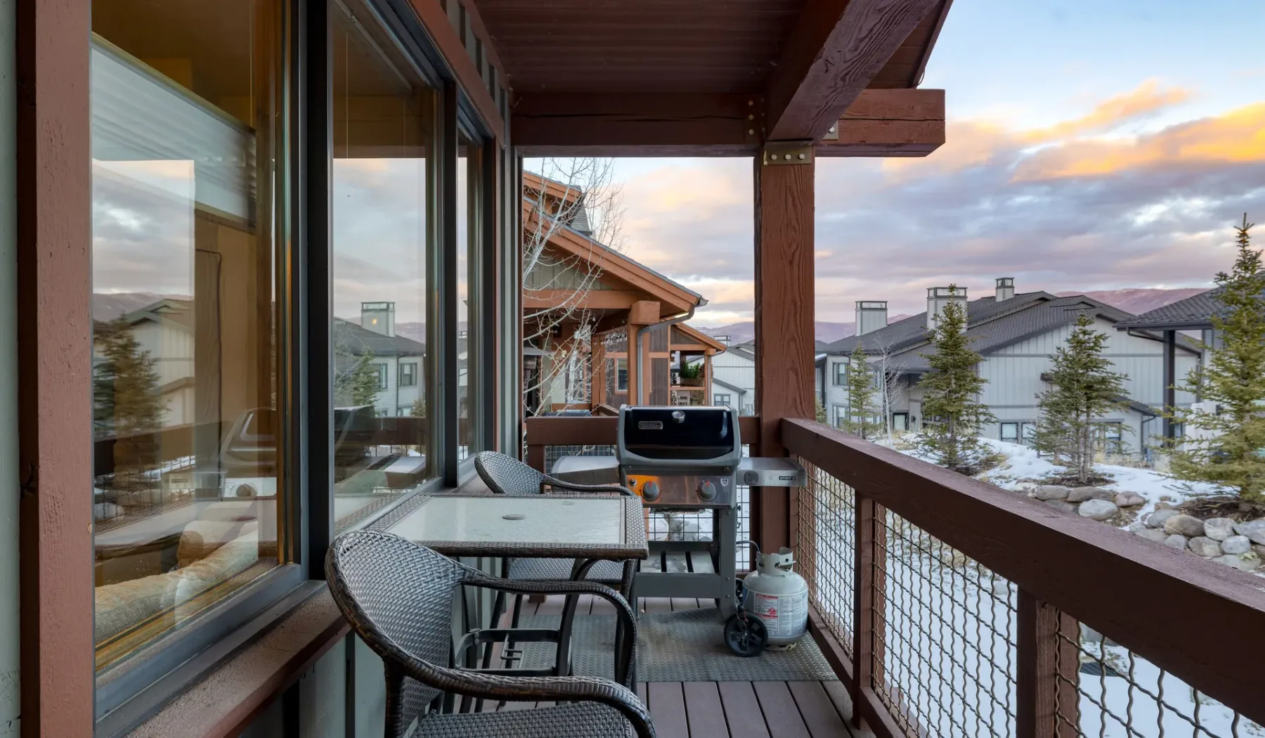 A covered outdoor deck with seating and a grill overlooks snow-covered homes at sunset.