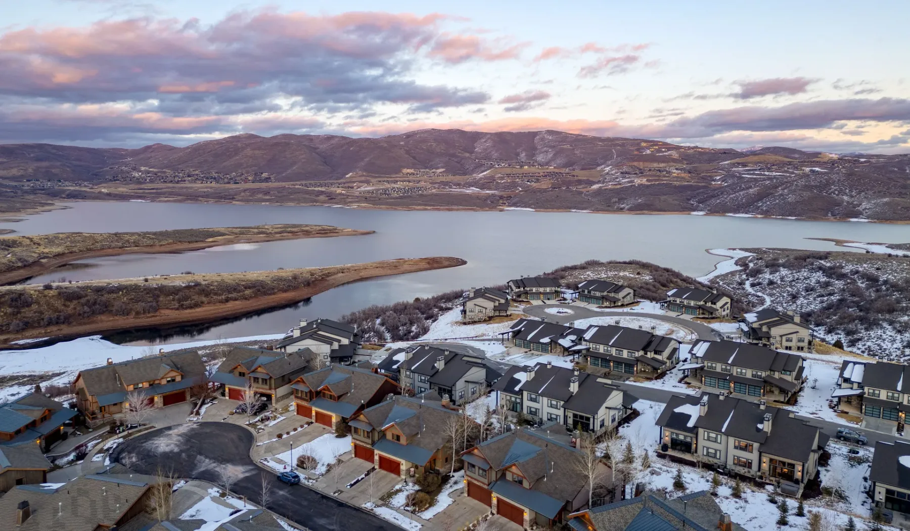 A snowy lakeside residential community sits below rolling hills at sunset.