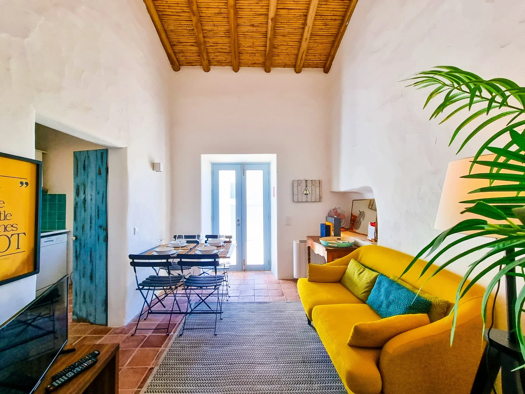 Un salón luminoso tiene un sofá amarillo, una mesa de comedor y un techo con vigas.