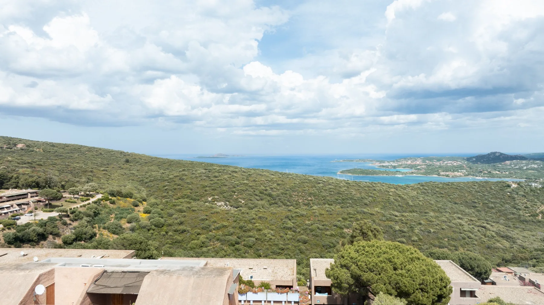 Un amplio paisaje costero con colinas verdes, edificios dispersos y el mar al fondo.