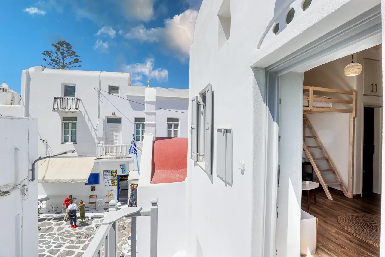 A white building overlooks a stone-paved courtyard, with a bunk bed visible through an open doorway.