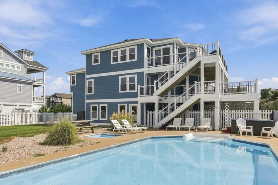A swimming pool sits beside a blue multi-story house with exterior stairs.