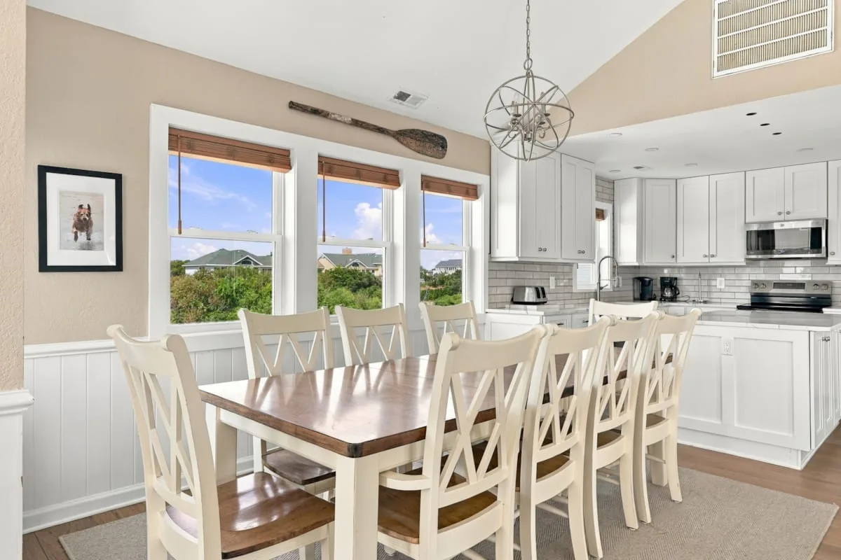 A dining area with a long table and white seating beside a bright kitchen.