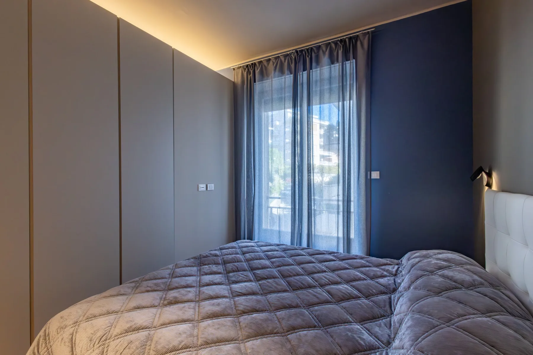 A bedroom with a quilted bed beside a tall window with sheer curtains.