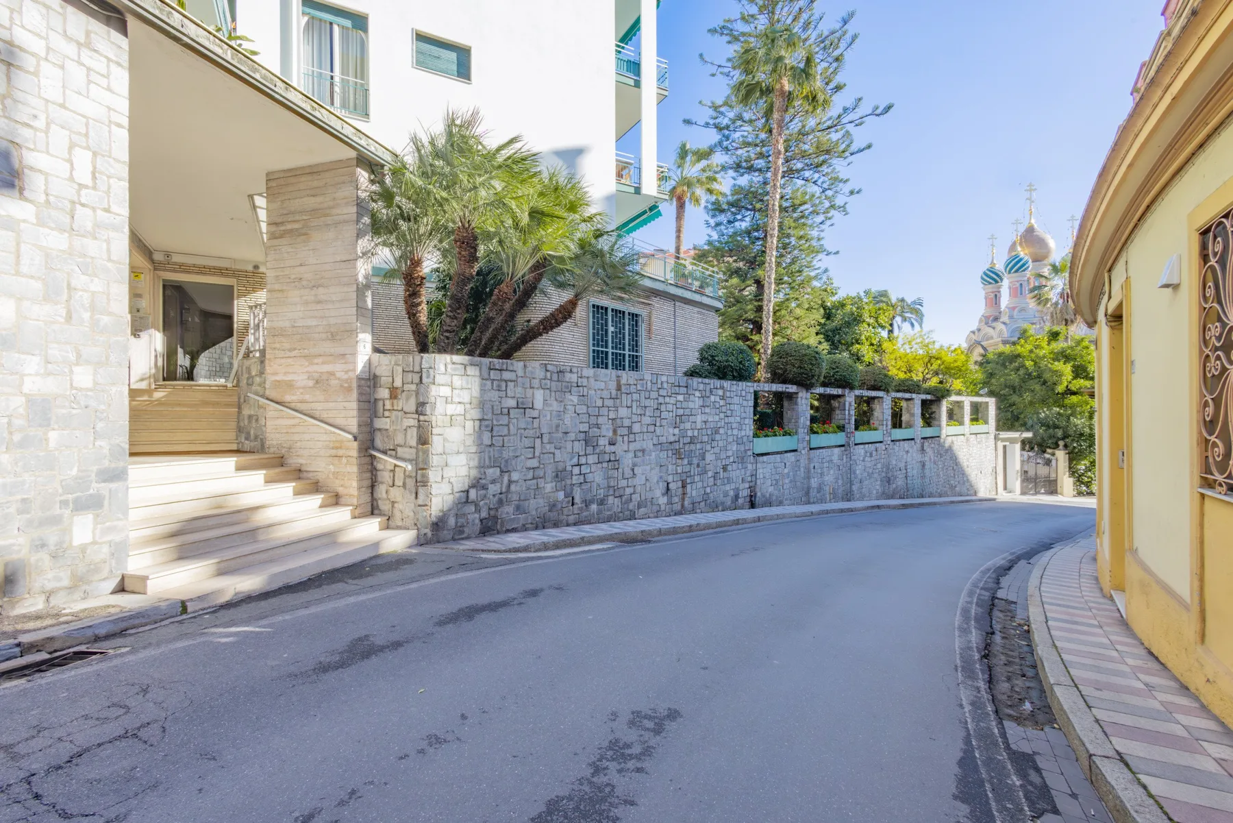 A curved street passes between stone walls and buildings, with trees and domed towers in the distance.