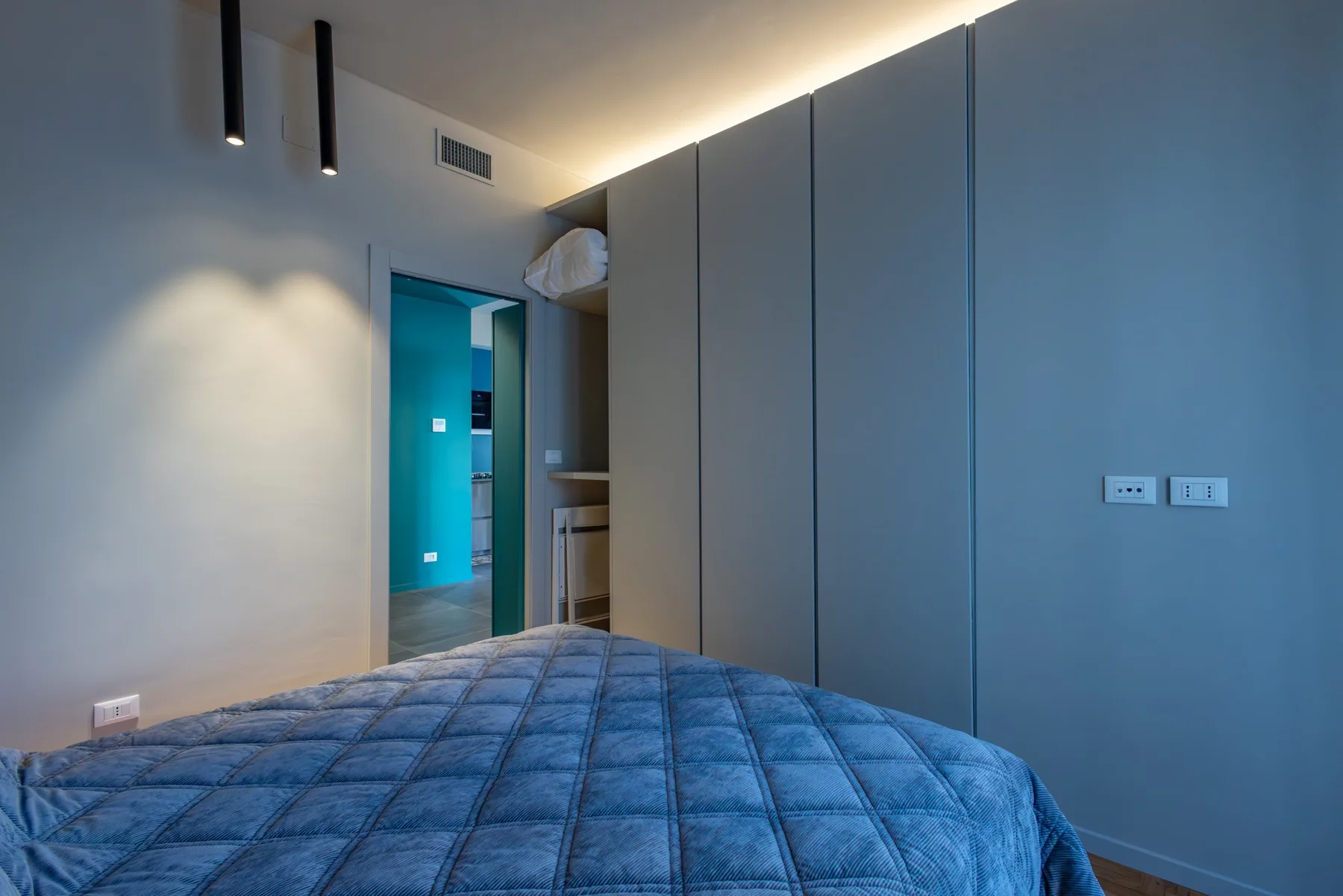A bedroom with a blue quilted bed facing a doorway to a brightly lit adjoining space.