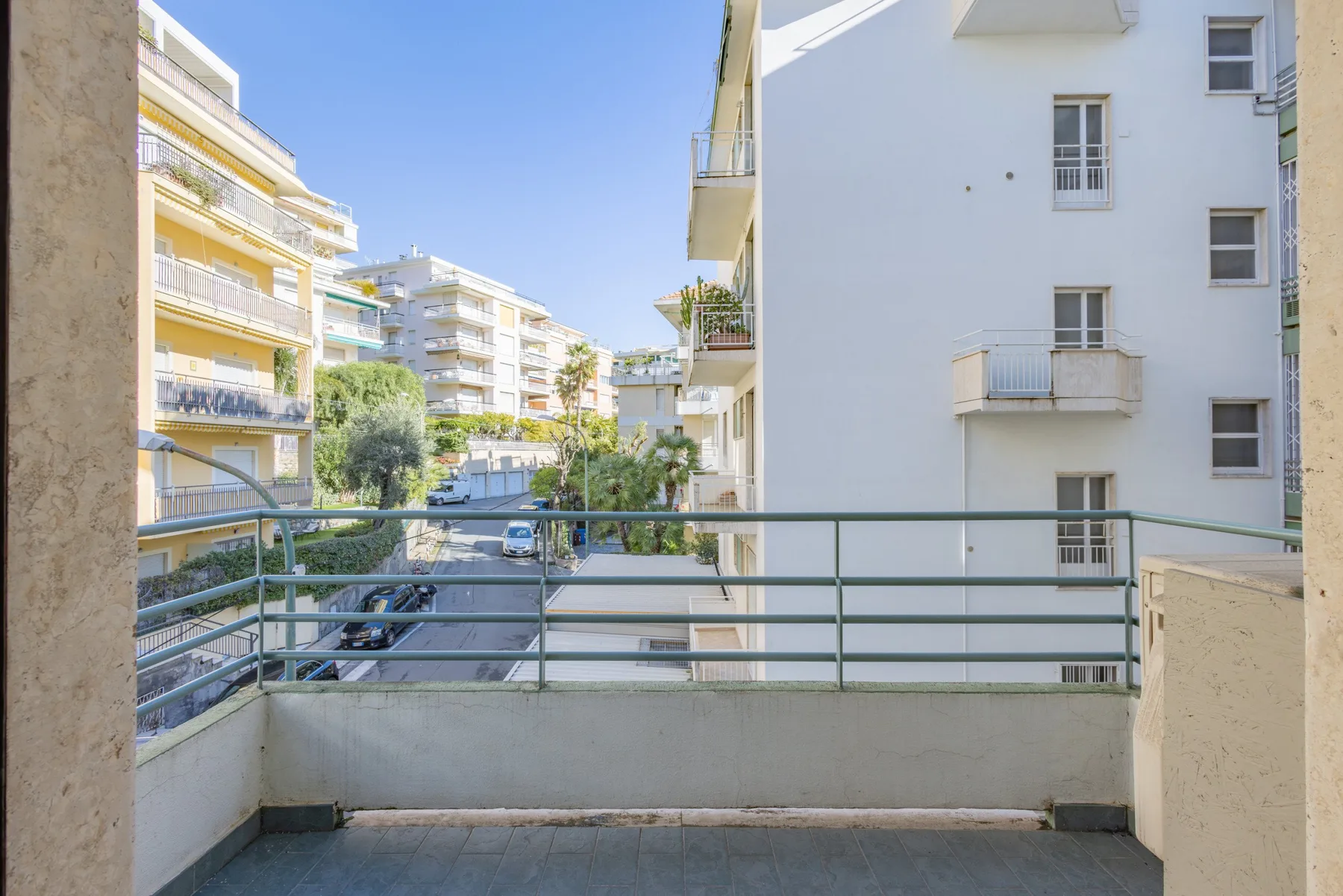 A balcony overlooks neighboring apartment buildings and a tree-lined street.