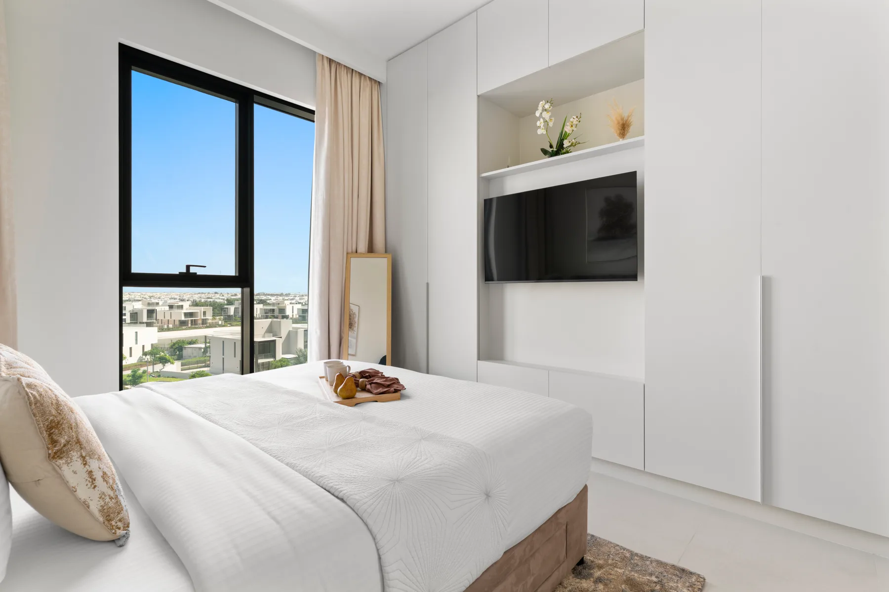A bright bedroom with a neatly made bed, large window, and wall-mounted television.