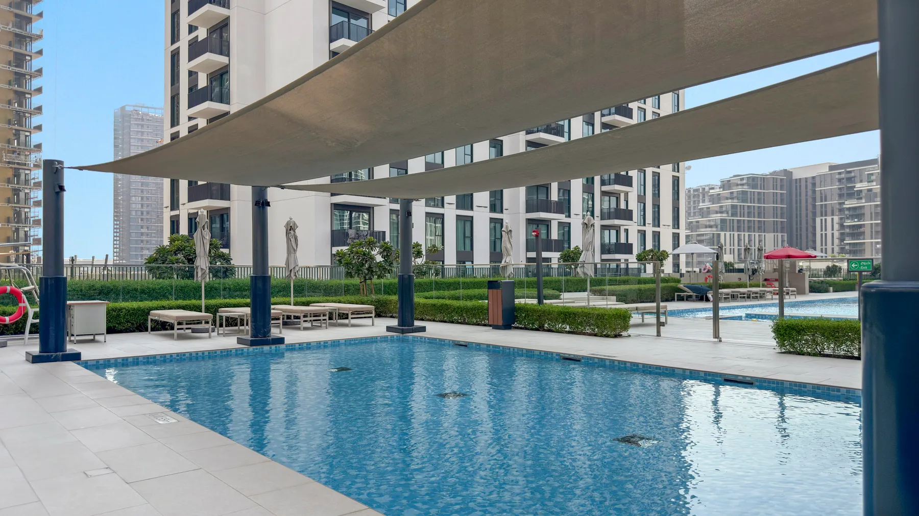 An outdoor swimming pool sits beneath shade canopies, surrounded by modern apartment buildings.