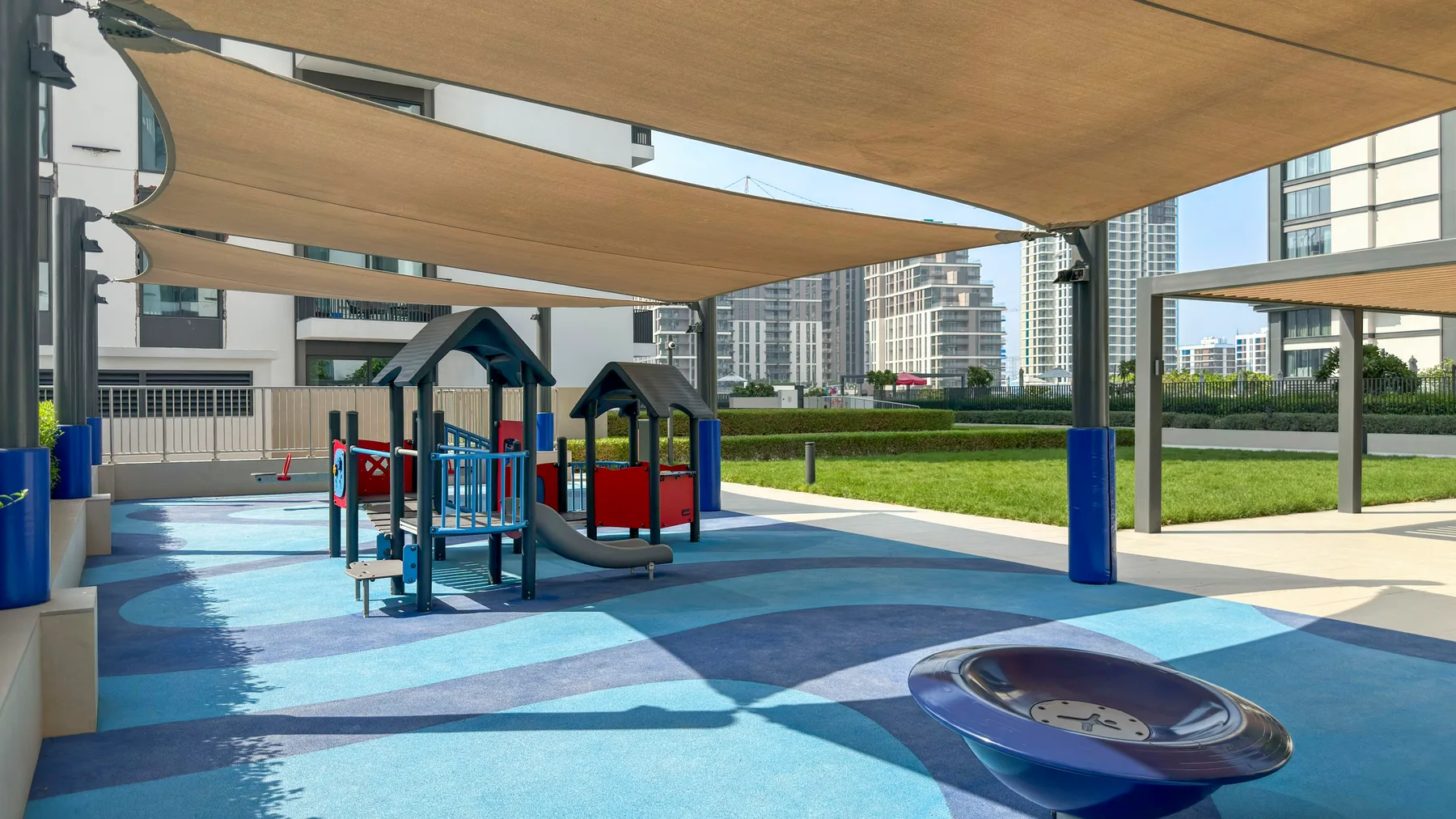 A shaded outdoor playground with colorful play structures and a rubberized surface.
