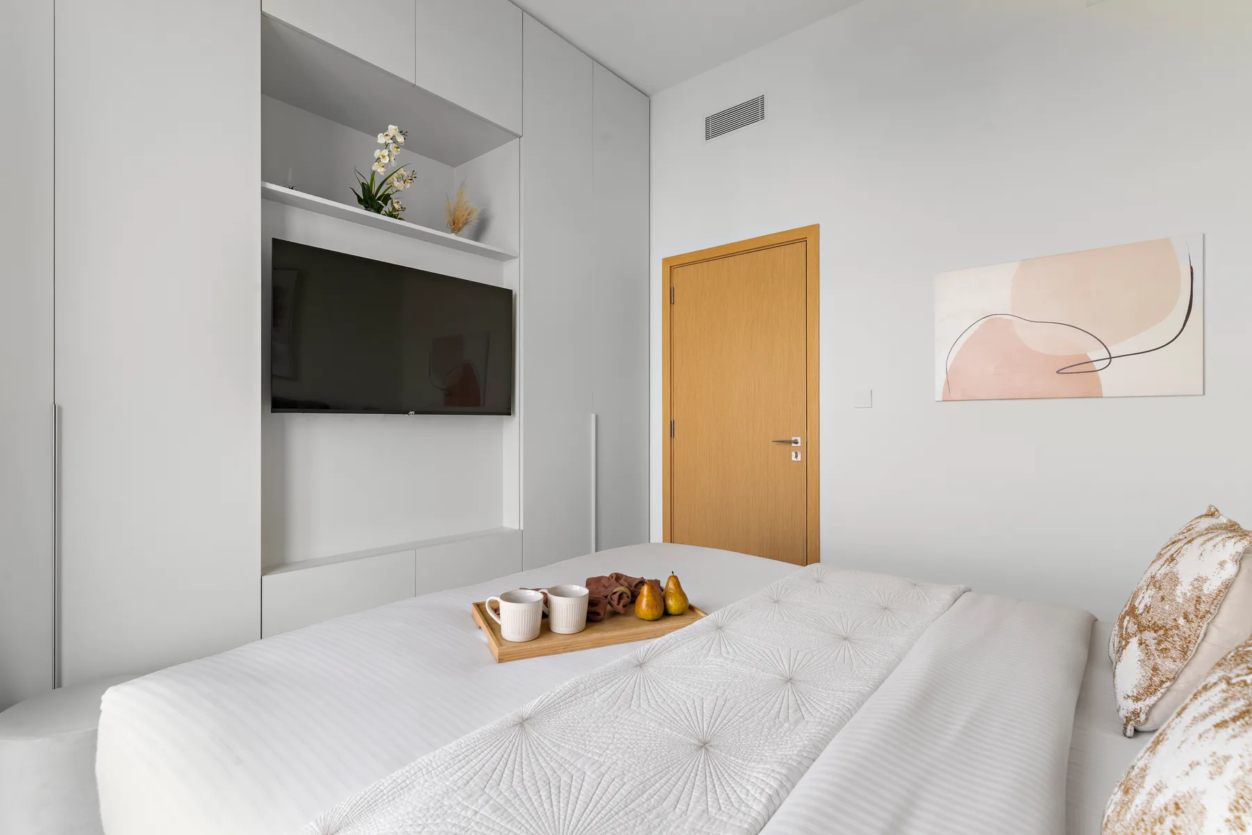 A bright bedroom with a neatly made bed, wall-mounted television, and abstract artwork.
