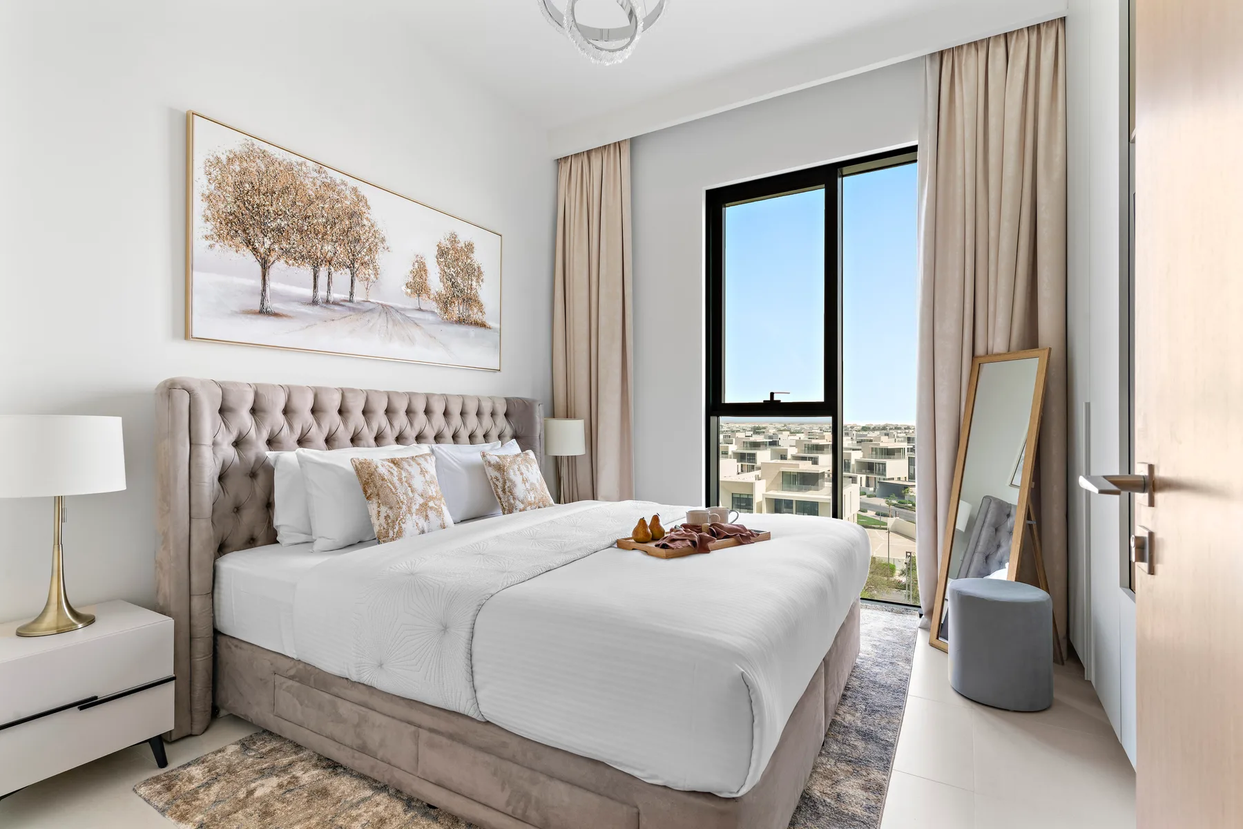 A bright bedroom with a large bed, neutral bedding, and a tall window.