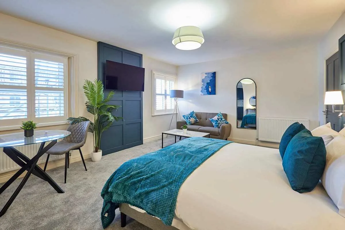 Un dormitorio con una cama, una manta verde azulado y asientos junto a las ventanas.