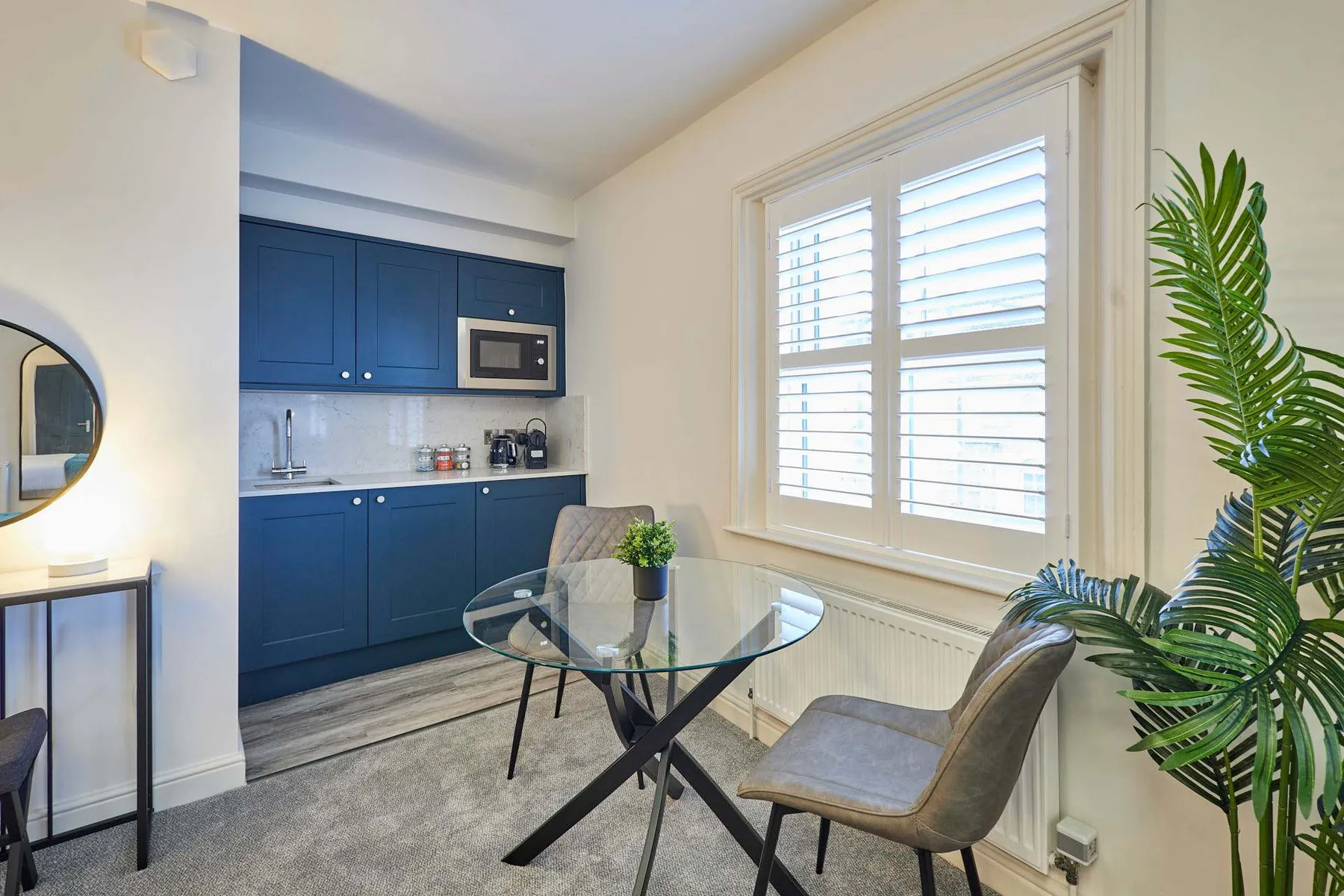Un comedor con una mesa de vidrio junto a una pequeña cocina azul y una ventana luminosa.