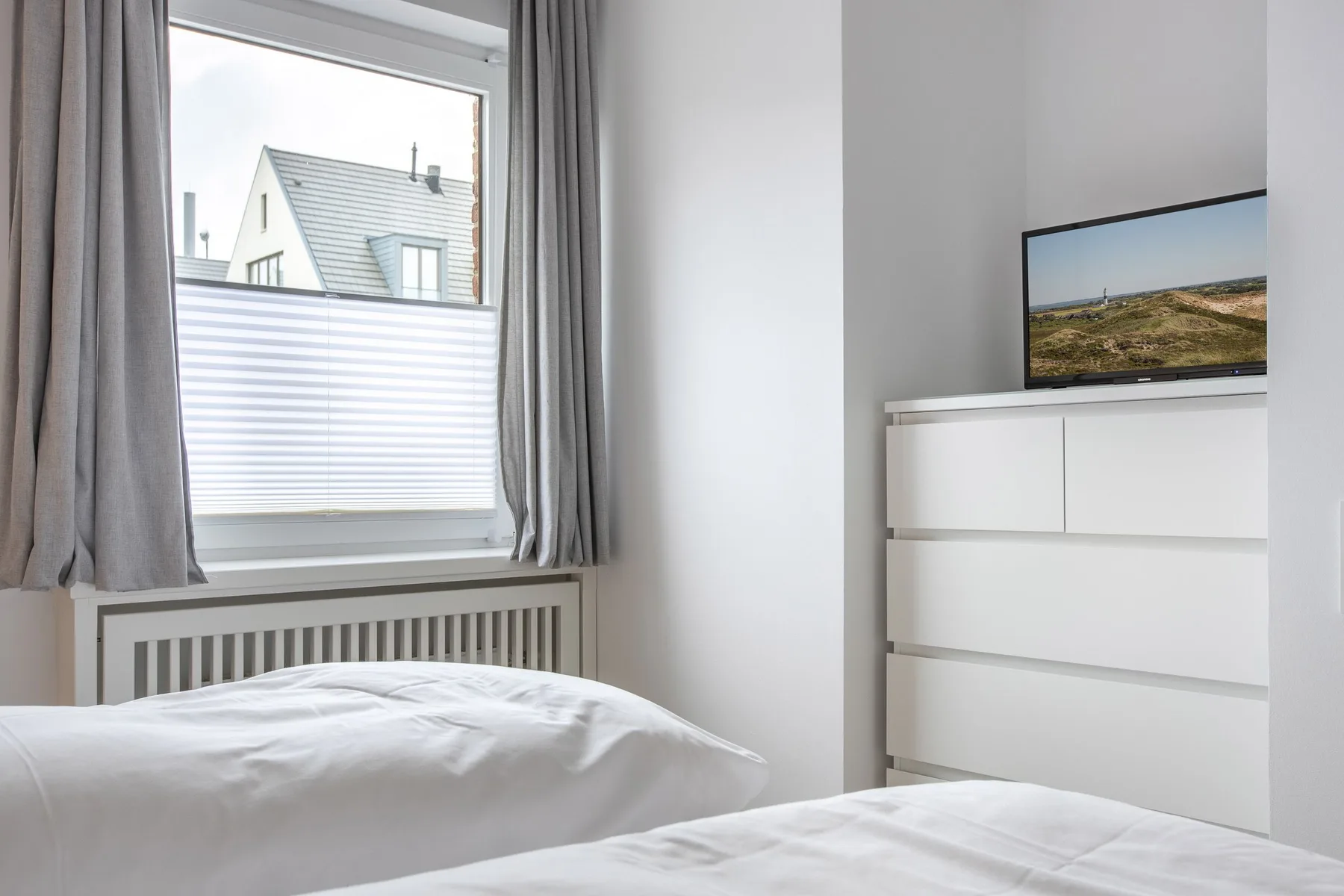 A bright bedroom with white bedding, a window, and a dresser topped by a television.