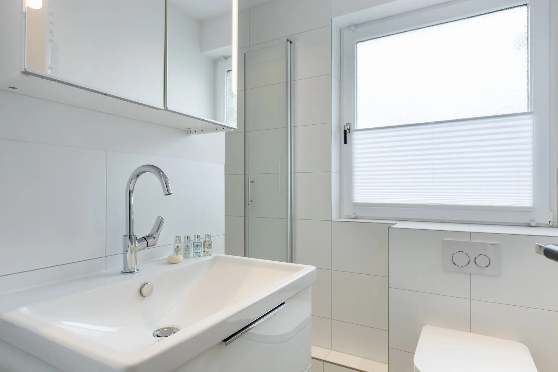 A bright bathroom with a sink, wall mirror, and frosted window.