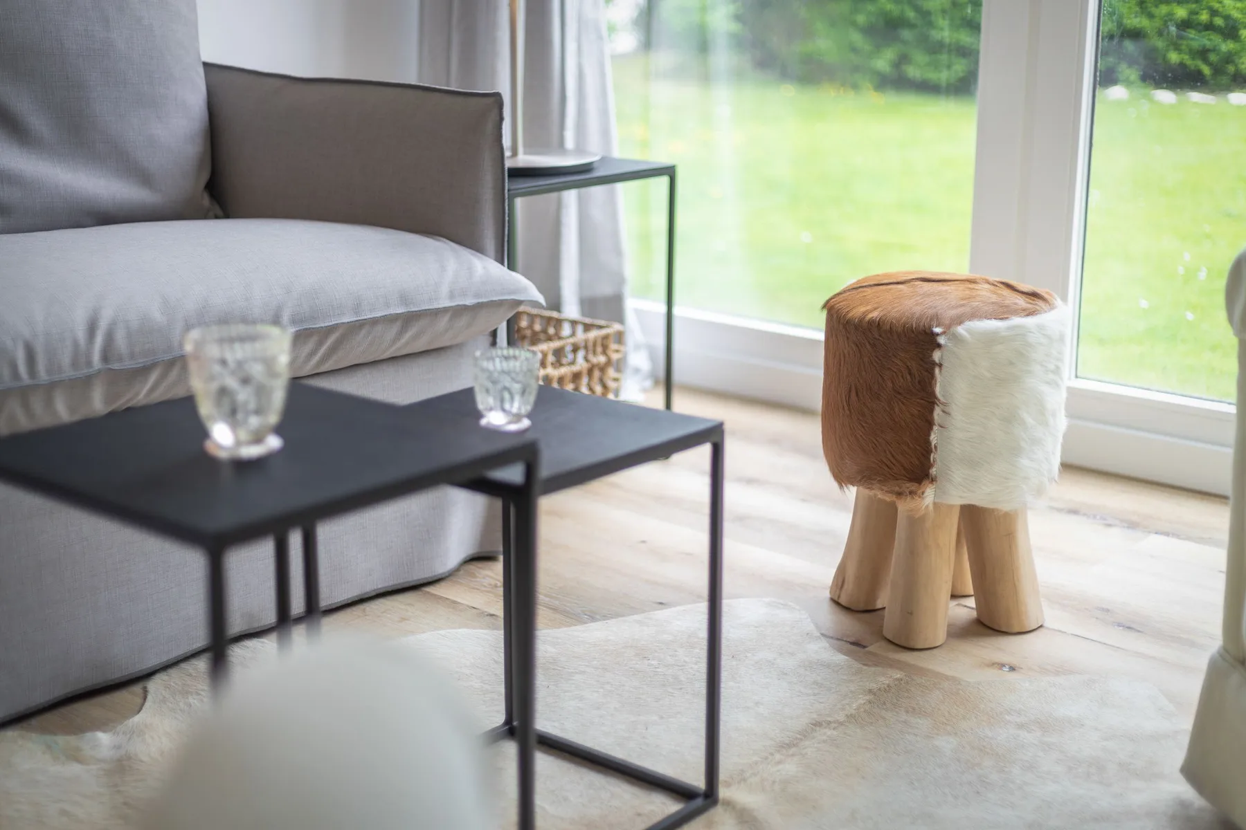 A living area with a gray sofa, low tables, and a small stool beside large windows.