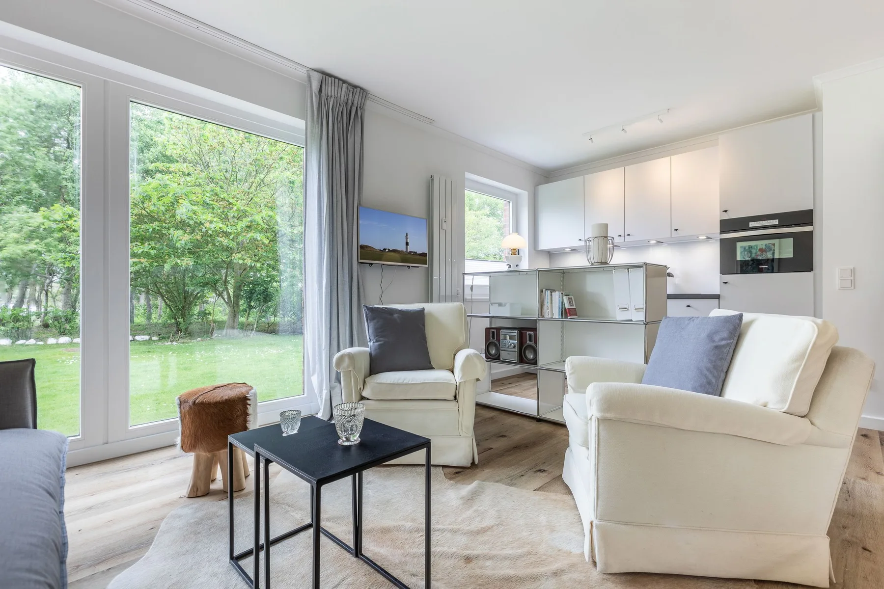 A bright living room opens onto a garden, with a compact kitchen behind the seating.