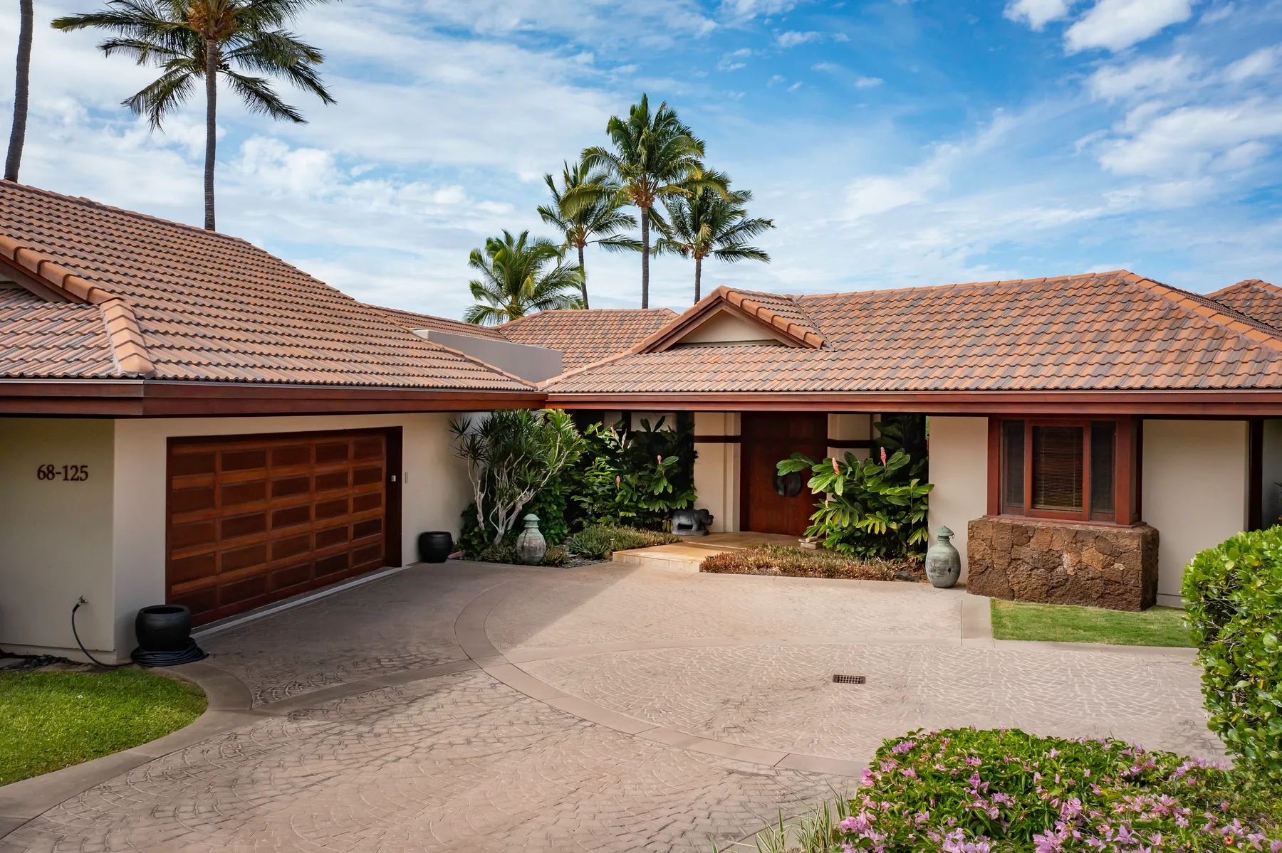 Exterior de una casa con una amplia entrada pavimentada, garaje y plantas tropicales.