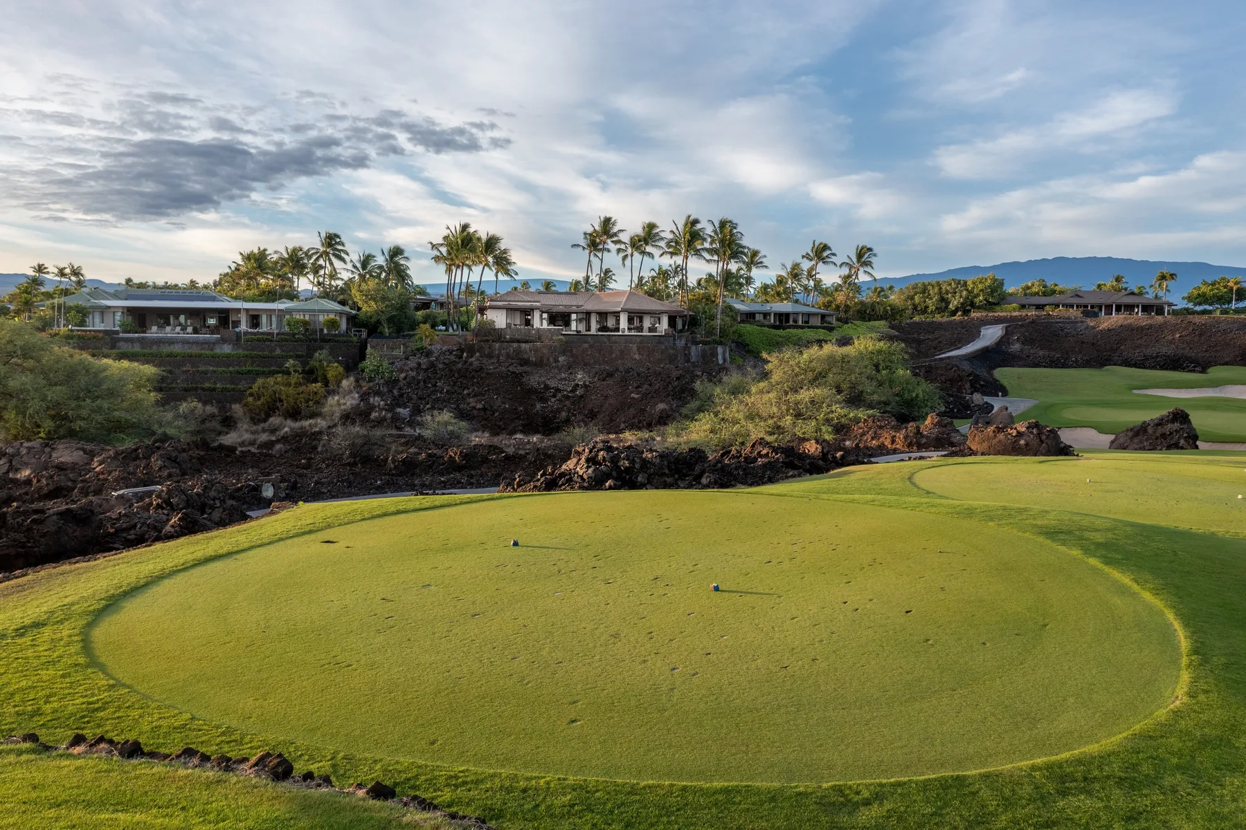 Un green de práctica circular está en un campo de golf ajardinado, con palmeras y edificios al fondo.