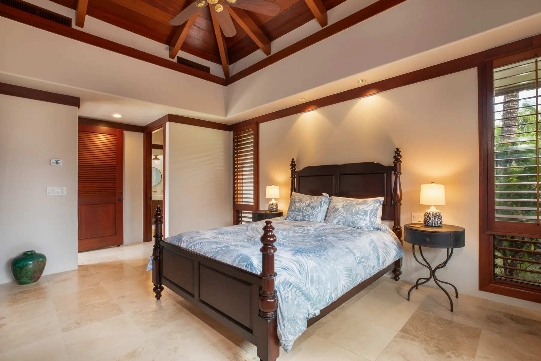 Un dormitorio con una cama de madera oscura, ropa de cama azul estampada y ventanas con persianas de madera.