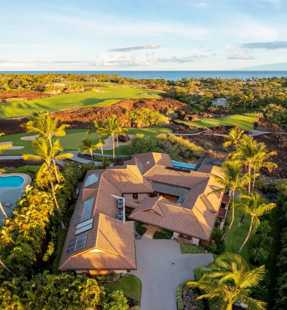 Vista aérea de una casa grande rodeada de vegetación tropical, con un campo de golf y el océano al fondo.