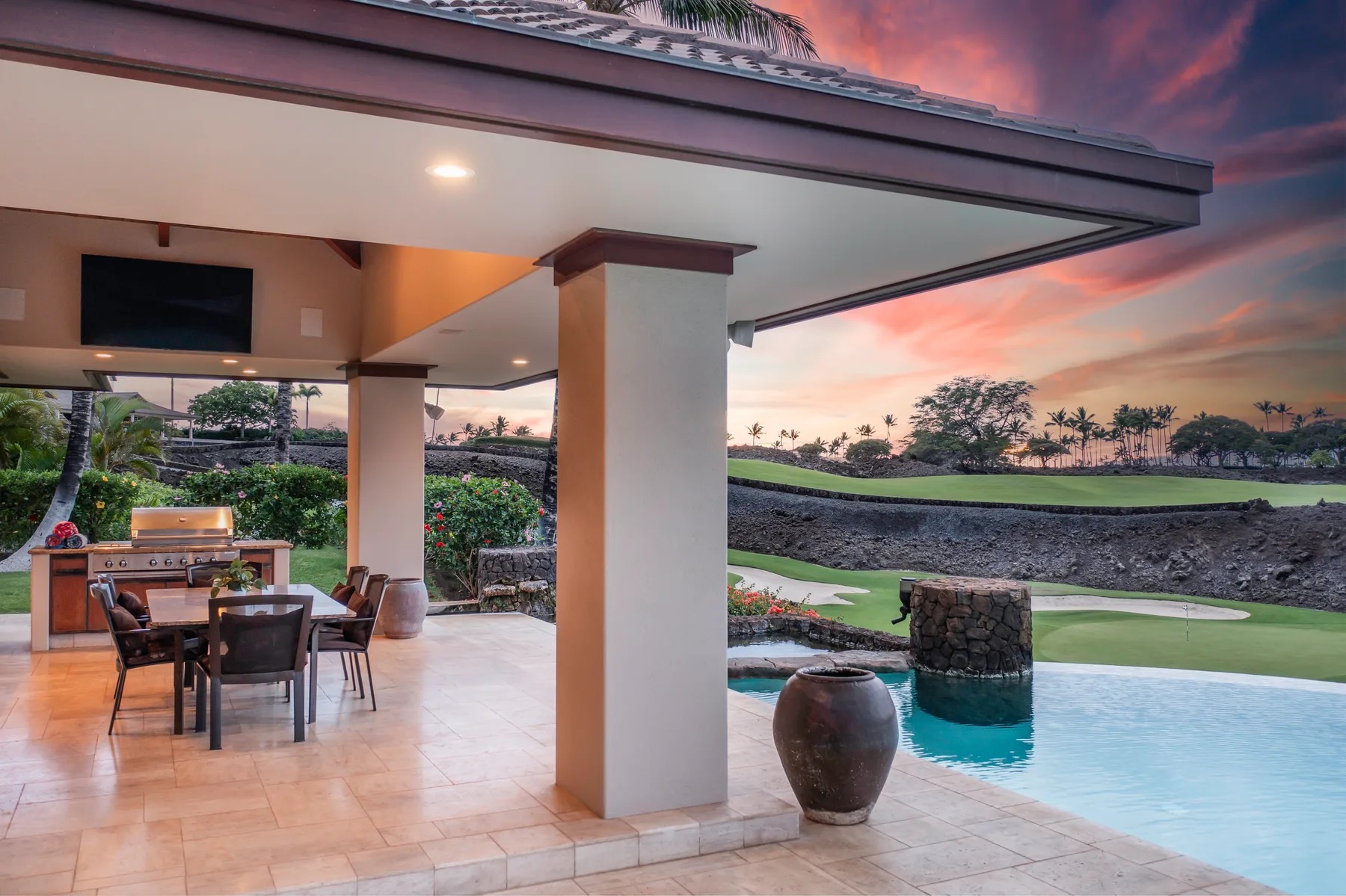 Un patio cubierto con asientos para comer da a una piscina y jardines al atardecer.