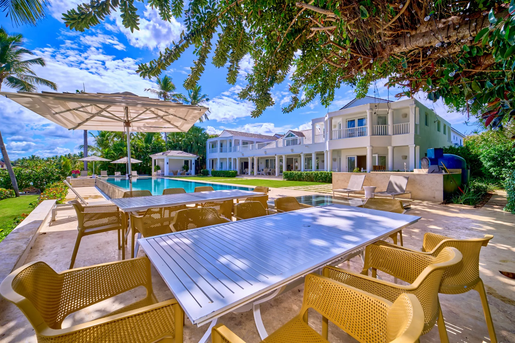 An outdoor dining area with tables and seating beside a swimming pool and white buildings.