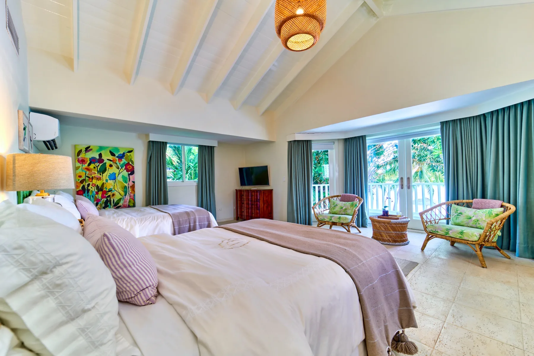 A bright bedroom with beds, colorful artwork, and a seating area beside large windows.