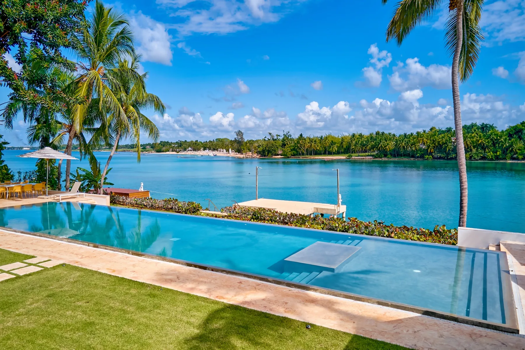 An outdoor swimming pool overlooks a calm blue bay lined with palm trees.